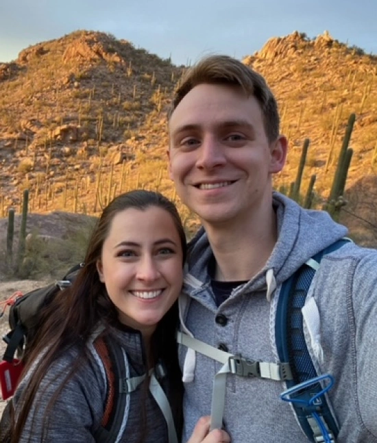 Two smiling people on a hiking trail with desert mountains and saguaro cacti in background.