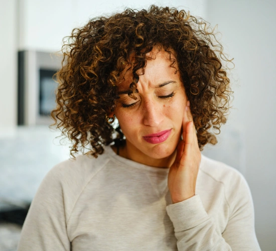Woman with curly hair touching face, wearing beige sweater, indoors, appears thoughtful or experiencing discomfort.