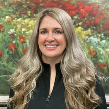 Woman smiling at camera with long blonde wavy hair, wearing black top, colorful flower garden background.
